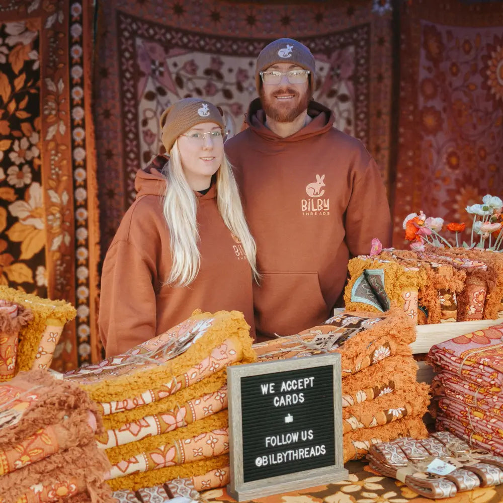Two people wearing brown hoodies standing behind a table with fabric and a sign that says 'We Accept Cards, Follow Us @BILBYTHREADS' against a patterned wall of woven picnic rugs.