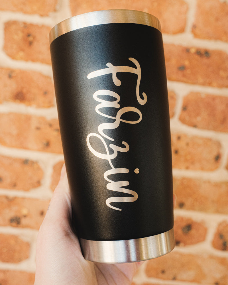 Black tumbler with 'Farzin' engraving held against a brick wall.