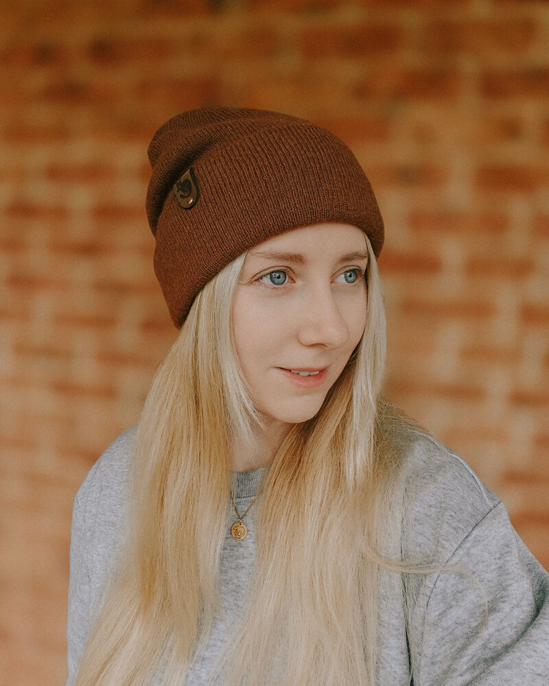 Person wearing a brown beanie and gray sweater against a warm-toned brick wall.